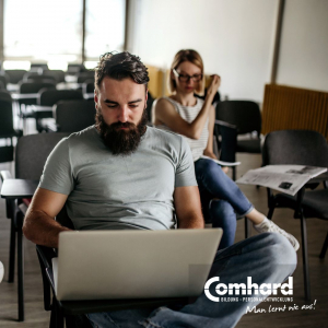 Ein Mann sitzt auf einem Stuhl mit einem Laptop auf dem Schoß. Eine Frau sitzt etwas versetzt hinter ihm