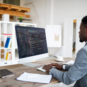 Mann sitzt vor einem Computer. Auf dem Display ist Coding Software zu sehen